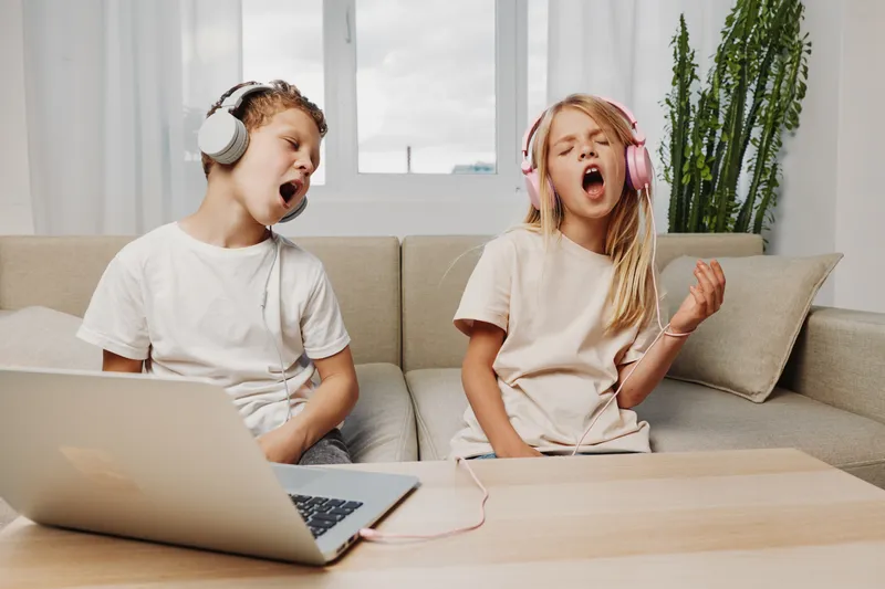 Two children wearing headphones sing passionately while sitting on a couch at home in casual clothes. The scene shows fun, creativity, music, and childhood entertainment indoors.