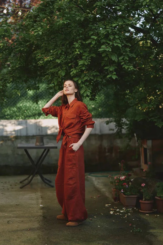 A young woman in a red jumpsuit stands in a leafy garden, adjusting her hair and striking a confident pose under dappled sunlight, blending relaxed style with bold color