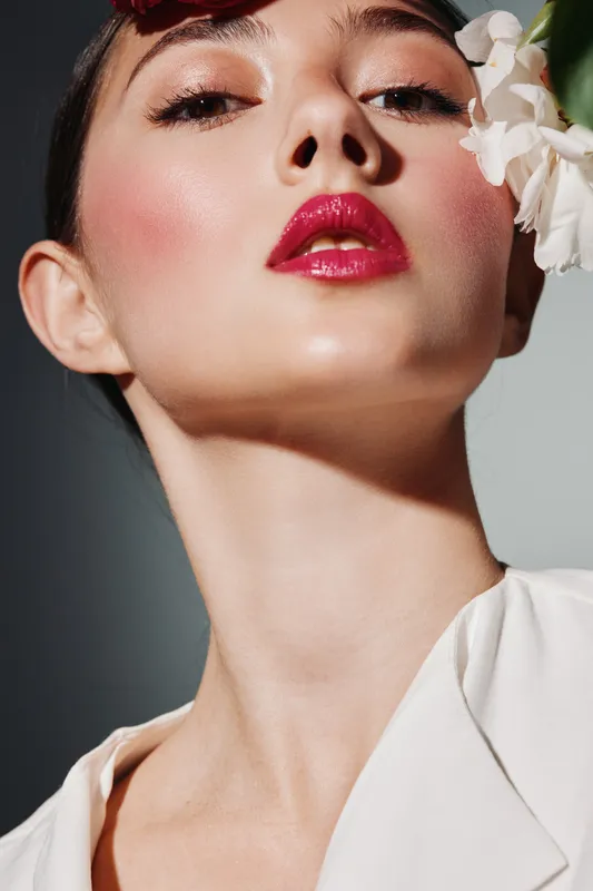Close-up beauty portrait of woman with glossy lipstick and dewy skin posing feminine calm with white flowers on dark background. Concept of fashion makeup skincare elegance.