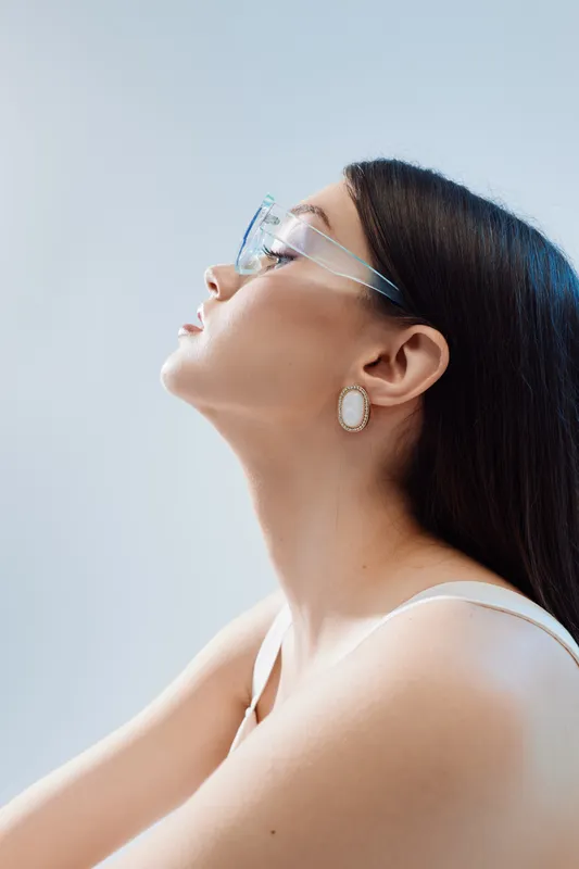 Profile of a young woman wearing transparent glasses and elegant earrings. She looks upward with closed eyes against a soft blue background. Concept of beauty fashion and confidence.