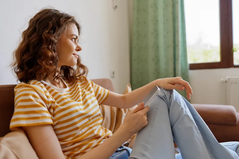 Woman relaxing at home sits on a couch wearing a striped shirt, casual jeans, gentle smile, natural window light, cozy living room scene, everyday lifestyle moment.