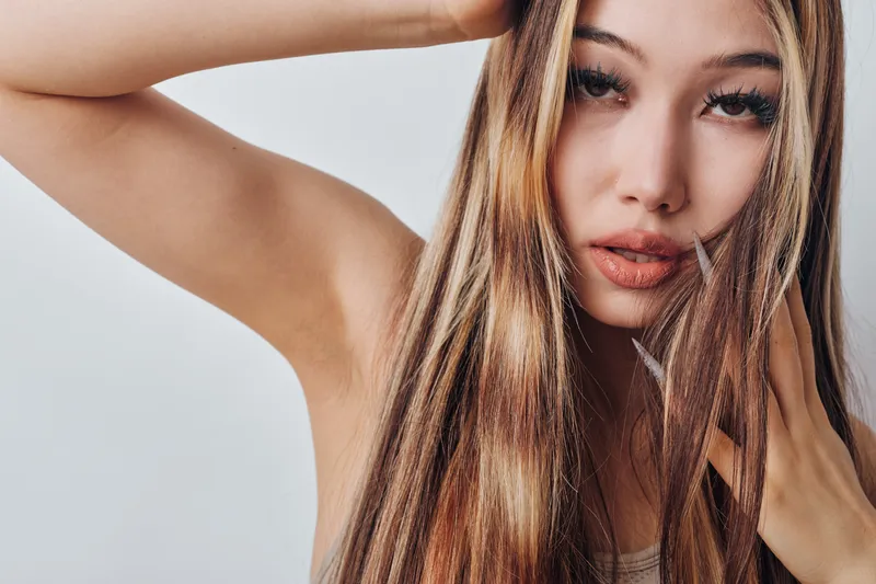 Young woman with long hair posing indoors with hand near face, natural makeup and serious expression on plain background with soft lighting.
