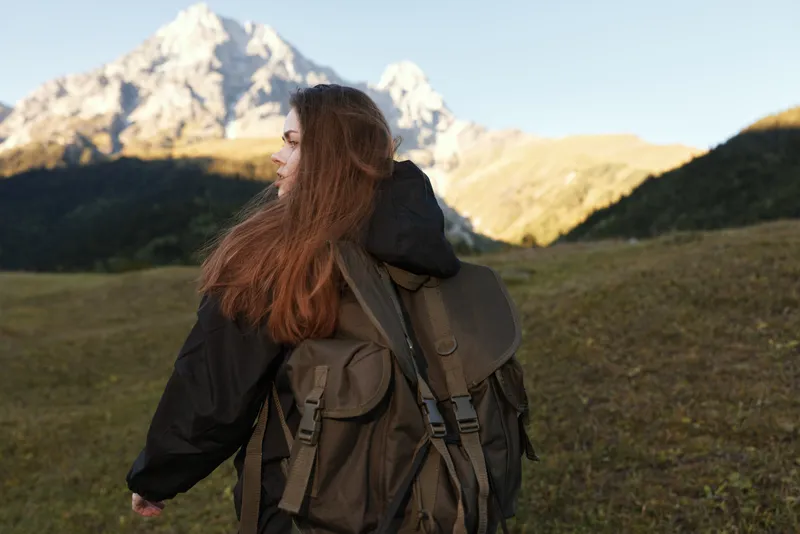 A lone hiker with a large backpack stands in a valley as rugged mountains rise in the distance during golden hour. The scene conveys adventure and exploration and a sense of vast wilderness.