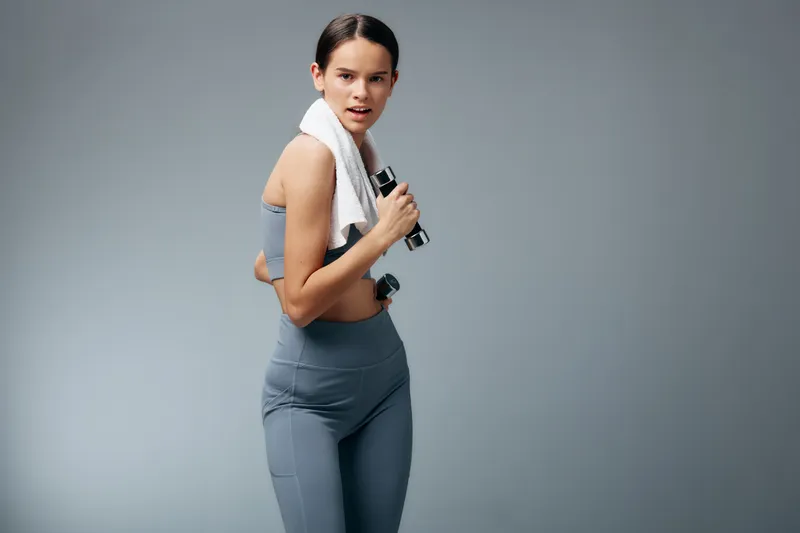 Fitness woman in athletic wear, holding dumbbells with a towel draped over her shoulder, mid workout pose in a studio. Strength, focus and endurance captured during a demanding training session.