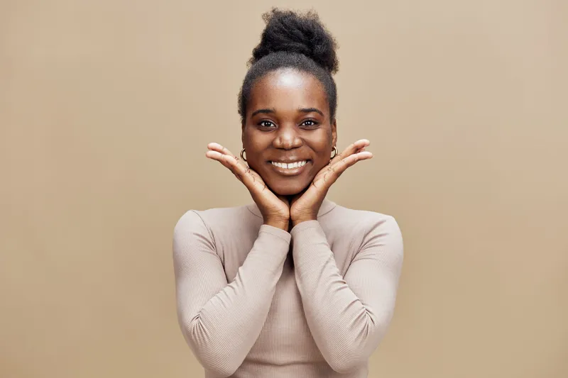 Smiling young Black woman with hands on her cheeks showcasing joy in a neutral toned background, perfect for representing happiness and positivity