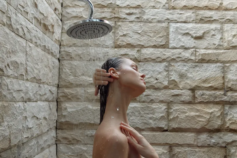 young woman enjoying a refreshing shower in a natural stone outdoor bathroom, water droplets on skin, peaceful and relaxed mood