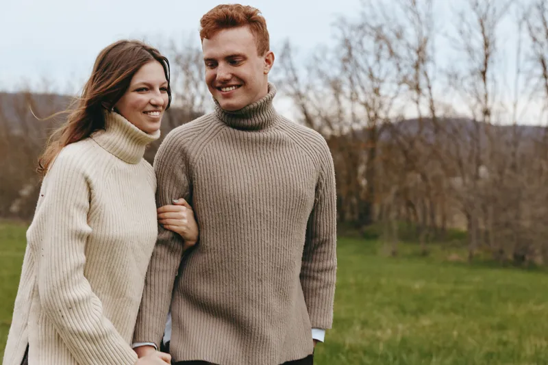 A young couple in knitted sweaters walks arm in arm through a grassy field during autumn, smiling and enjoying a peaceful outdoor moment together.