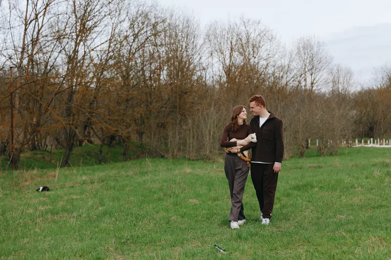 Loving couple outdoors in a park with a goat kid in arms, sunlit grass and bare trees in the background, lifestyle moment showcasing affection, companionship, casual fashion, and a gentle, wholesome