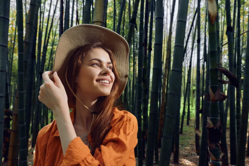 Woman smile hat bamboo forest portrait of a young traveler in nature, wearing straw hat and orange shirt, looking joyful among tall green stalks, outdoor lifestyle and eco travel.