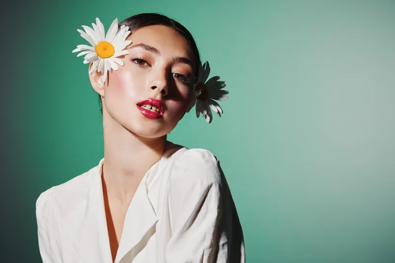 Portrait of a young woman with fresh makeup and daisies in her hair wearing white. Green background setting creates a fashion beauty concept with natural creativity and soft light.