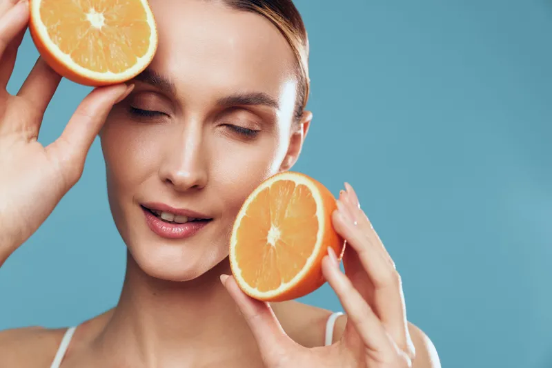 Woman skincare spa, orange citrus face beauty, relaxation with closed eyes, holding fresh orange slices near cheeks, gentle hand touch, blue background, clean minimal aesthetic