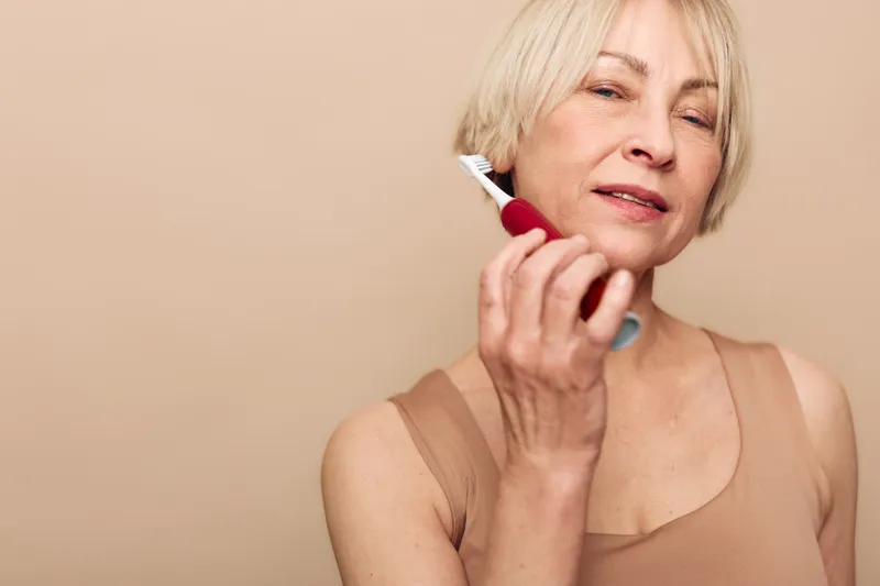 Mature woman with blonde hair using electric toothbrush in beige tank top. Skincare and oral hygiene concept with natural light and neutral background for health and self-care.