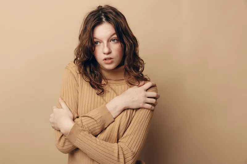 Young woman with curly hair wearing beige sweater looks worried and hugs herself. Neutral background and soft lighting create a mood of anxiety and introspection.