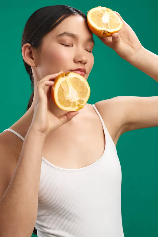 asian woman wearing a light top holds fresh orange halves near her face, calm and playful expression, vibrant citrus tones against a teal background, modern lifestyle feel.