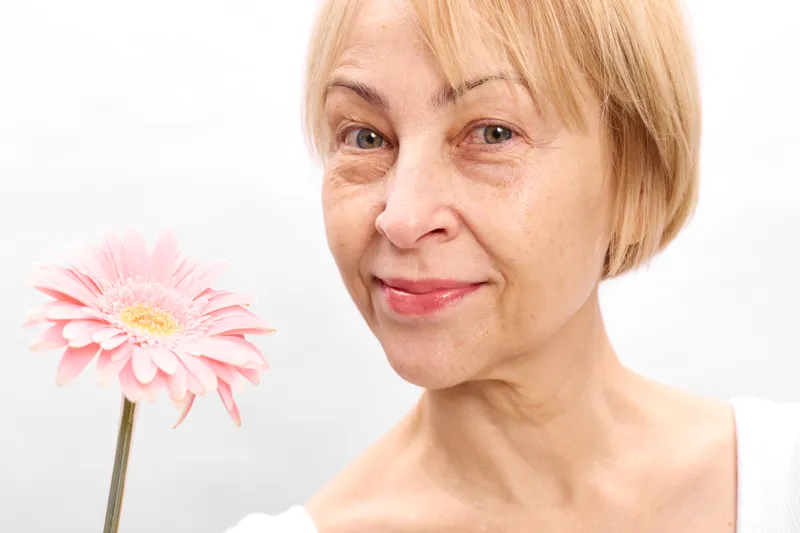 Woman with blonde hair in close up portrait holding pink flower, studio lighting, gentle smile, beauty and skincare concept, clean white background, calm serene mood