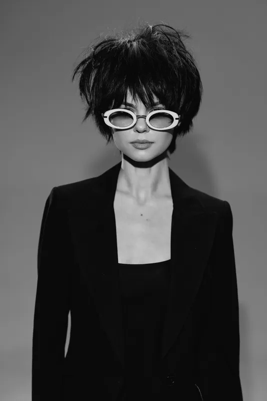 Stylish black and white portrait of a woman wearing unique sunglasses and a dark blazer with an edgy hairstyle.