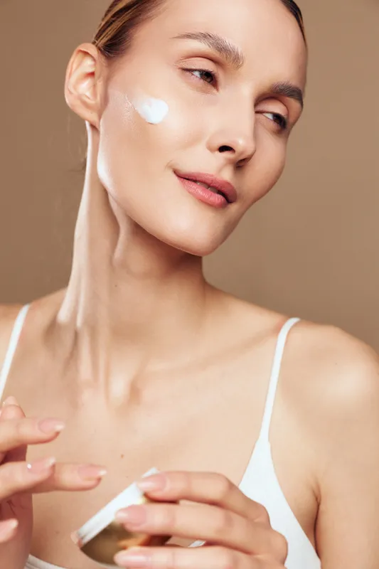 A young woman with clear skin applies moisturizing cream to her cheek in a close up beauty portrait. The neutral background highlights skincare and self care concepts.