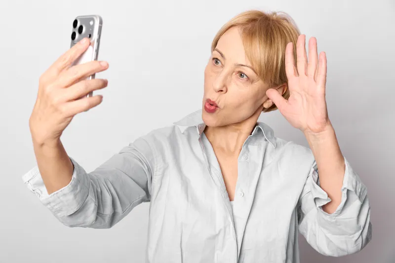 Surprised woman selfie smartphone touching ear, close portrait, soft studio lighting on white background, beauty skincare self care concept, technology and digital communication