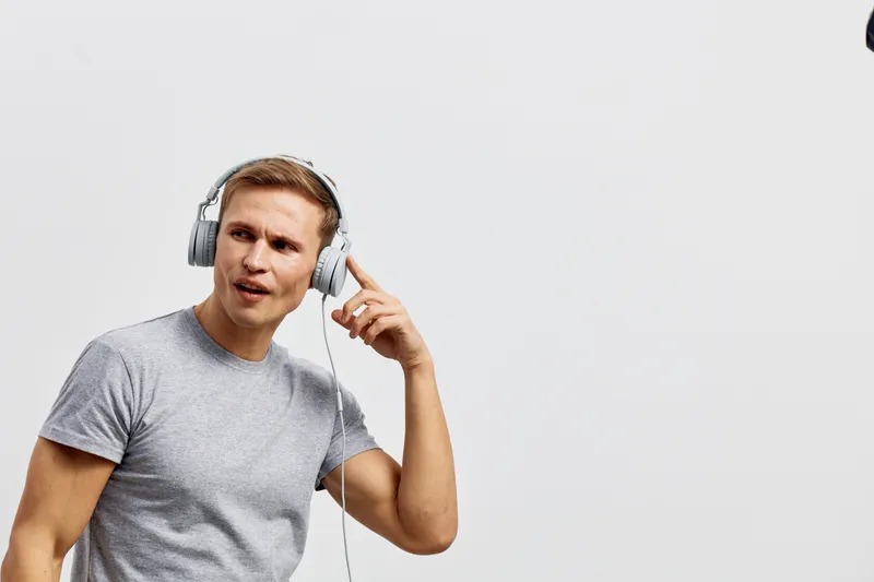 Young man in casual gray T-shirt wearing large headphones, listening to music with expressive face, isolated on plain white background. Studio portrait of a guy enjoying audio entertainment. Lifestyle