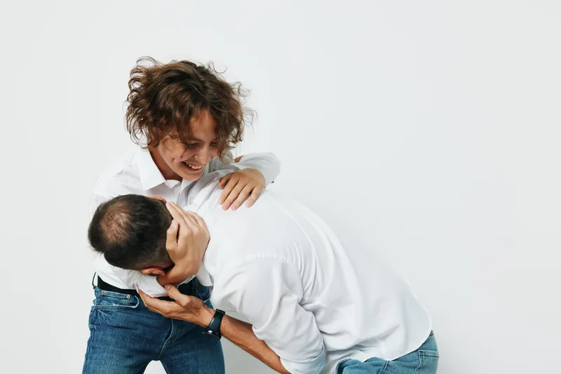 Professional man stands against a plain colored backdrop, showing strong emotion as a colleague playfully engages in a light interaction, conveying energy, confidence, and teamwork in a studio