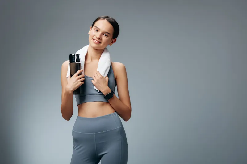 Fitness woman in athletic wear with towel over shoulders and water bottle, posing confidently in a studio to showcase sport mindset and healthy lifestyle.