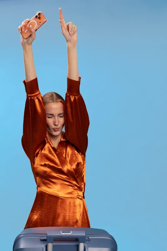 Young woman in shiny orange dress holds smartphone and raises arms, standing behind blue suitcase. Concept of travel, celebration, vacation, and lifestyle with joyful mood on blue background.