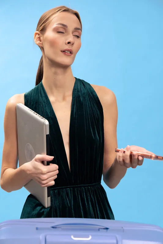 Confident woman in elegant dark green dress holds laptop and smartphone while standing behind blue suitcase on light blue background. Concept of business travel and modern technology.