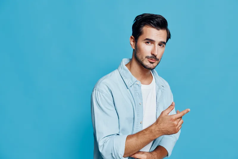 Stylish young man wearing light blue shirt on blue background pointing with finger, expressing confidence and creativity in a modern studio portrait.