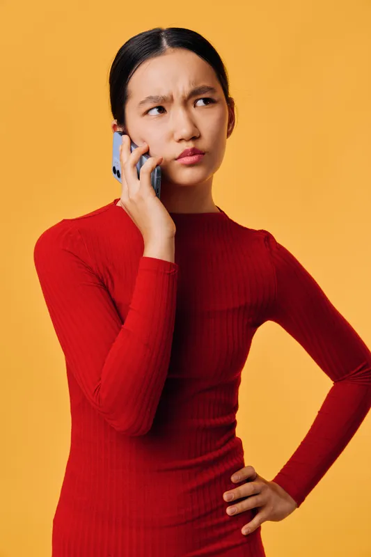 Young woman in red dress with hand on hip talking on smartphone with skeptical and confused expression against orange background. Female, communication, technology, emotion and modern lifestyle.