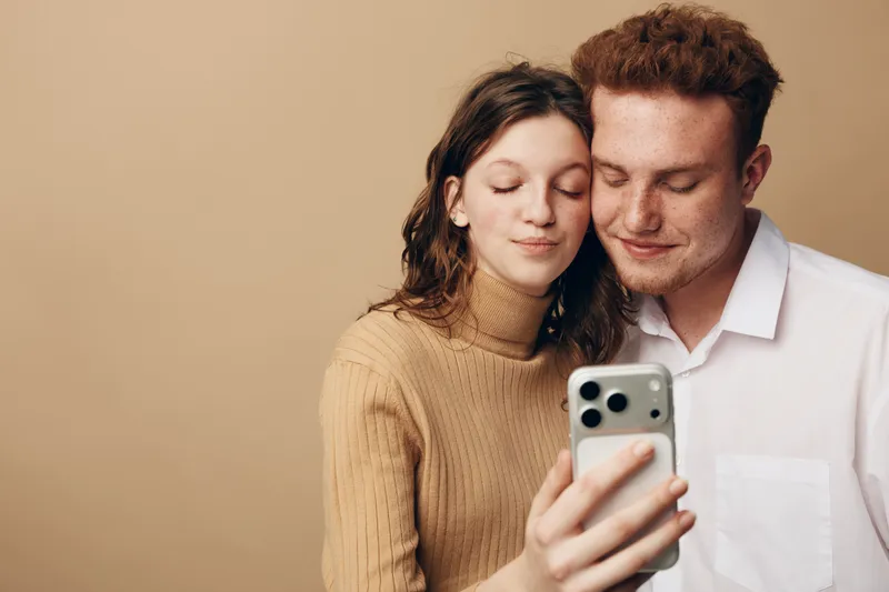 Young couple taking a selfie together with a smartphone. They smile gently with closed eyes, showing affection and closeness in a warm indoor setting with neutral background.