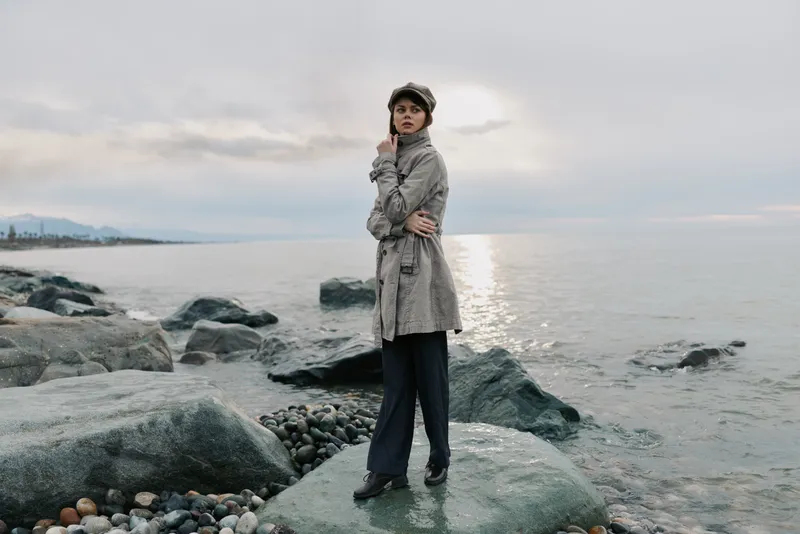Young woman in grey coat and hat stands on rocky shore near calm sea under cloudy sky. Moody outdoor scene with casual fashion and peaceful nature landscape at sunset or sunrise.