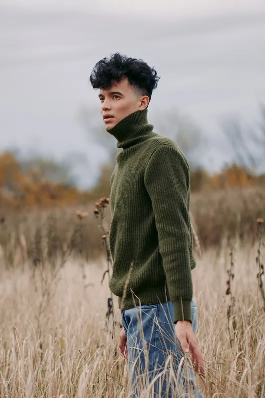 A solitary portrait of a young man in a green turtleneck sweater standing in a windswept field, exuding authenticity and credibility through relaxed posture and honest light.