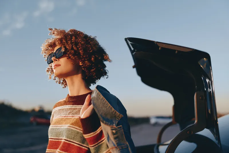 Woman lifestyle at sunset, outdoors, sunglasses on, curly hair, denim jacket and striped sweater, standing beside a car door, calm vacation travel, coastal road mood