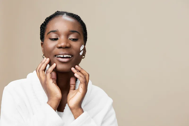 Young African woman applying skincare product, smiling happily in a soft white robe against a neutral beige background, embodying self care and beauty