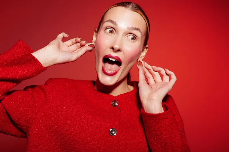 Excited woman in red sweater poses with open mouth and raised hands on red background. Concept of joy, surprise, fashion, and vibrant emotions in studio portrait photography.