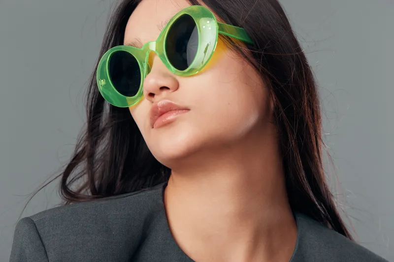 Woman wearing large green sunglasses and gray jacket posing with neutral expression and loose hair against plain studio background for fashion and style portrait.