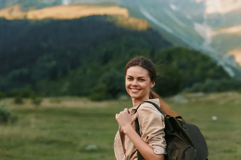A young woman with a backpack smiles as she hikes through a grassy field with distant mountains on a sunny day, conveying curiosity, independence, and adventure.