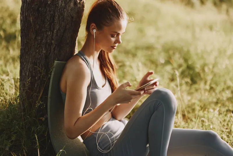 Young woman enjoying music on her smartphone while sitting outdoors, reflecting a healthy lifestyle and relaxation, with natural green background
