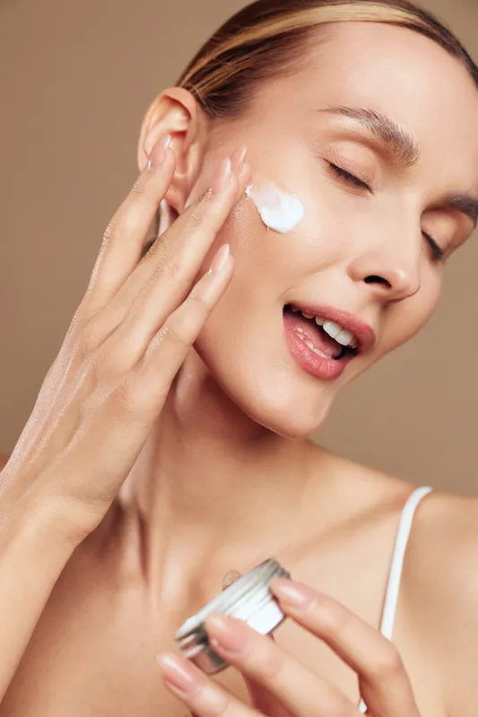 Young woman applying face cream with closed eyes, smooth skin, natural makeup, soft lighting, skincare routine, beauty care, close-up portrait, beige background