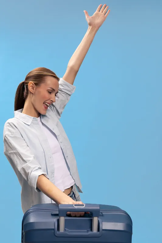 Happy woman with suitcase raises arm in excitement against blue background. Travel concept with casual clothing and joyful expression. Ready for vacation or trip.