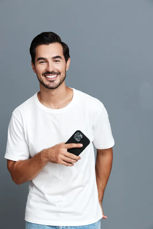 Happy smiling young man in casual white t-shirt holding smartphone while standing against a neutral gray background. People lifestyle concept.