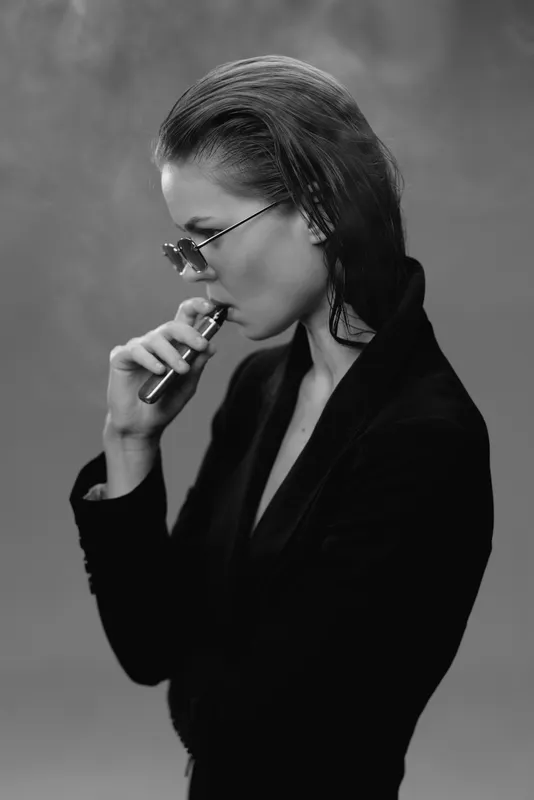 Black and white portrait of a thoughtful woman in sunglasses vaping with a dark background and stylish black outfit