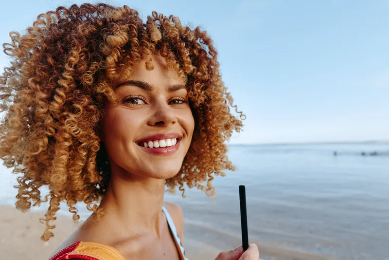 A happy young woman with curly hair smiles while holding a straw at the beach. The calm sea and clear sky create a peaceful summer atmosphere in the background.