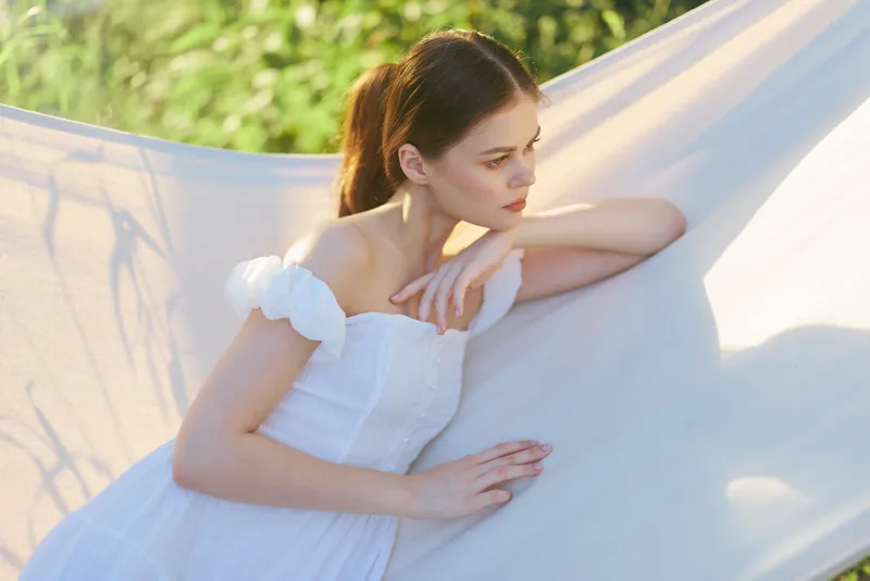 Young woman in a white dress resting against a flowing fabric backdrop in a sunlit outdoor setting, conveying serenity and peacefulness