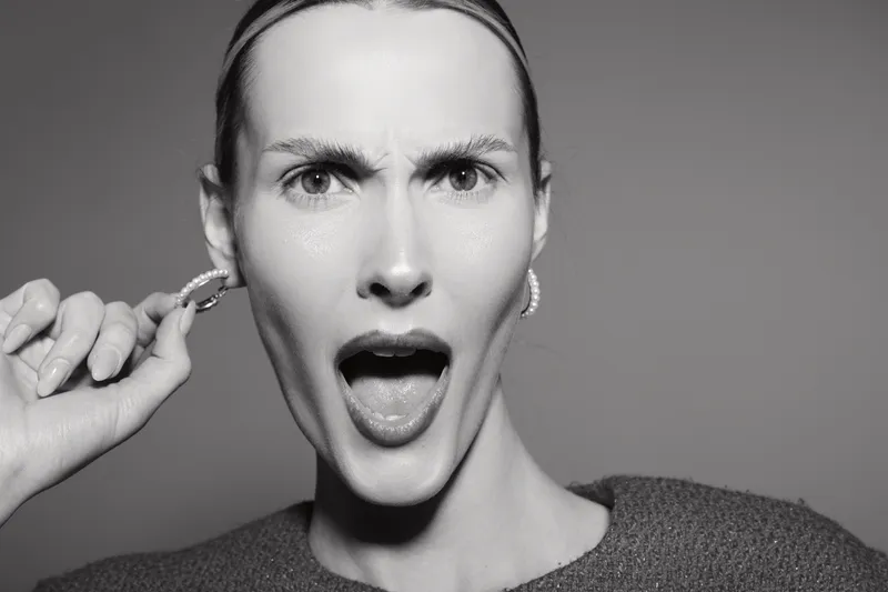 White woman with pale skin and slicked hair expresses surprise or shock holding hoop earring near ear in studio with plain background. Concept for emotion, gesture, and facial expression.