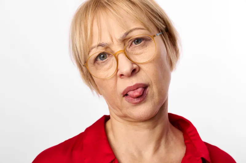 Elderly woman portrait, closeup face with blonde short hair, wearing glasses and red shirt, direct gaze toward camera, studio light, neutral white background, calm serious expression