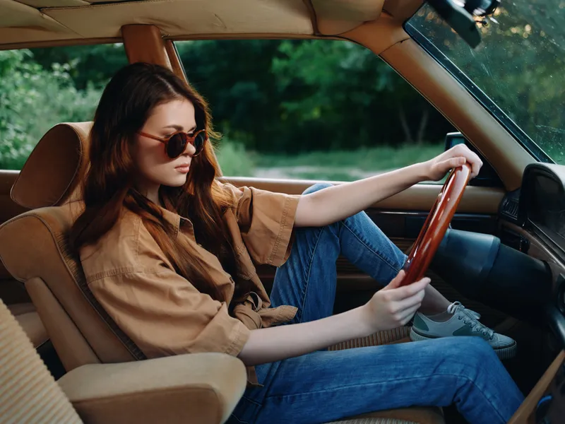 Young woman in casual attire driving a vintage car, displaying confidence and focus, surrounded by lush greenery, perfect for themes of freedom and adventure.