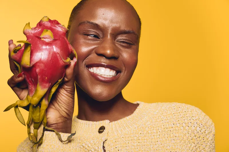 Smiling dark skinned African woman holds fresh dragon fruit close to her face with eyes closed, wearing beige sweater. Concept for healthy lifestyle, exotic fruit, and natural beauty.
