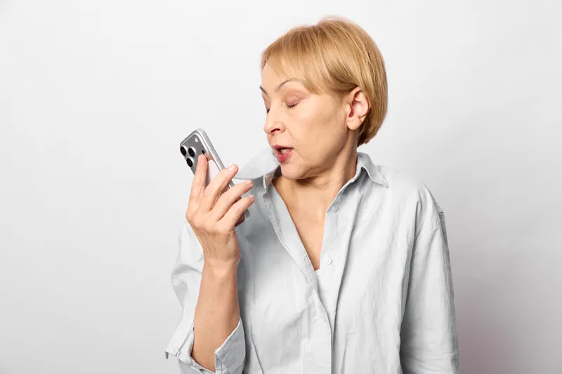 Elderly woman holding a smartphone near her face, talking, indoors studio portrait on a white background, technology communication moment, attentive expression soft light