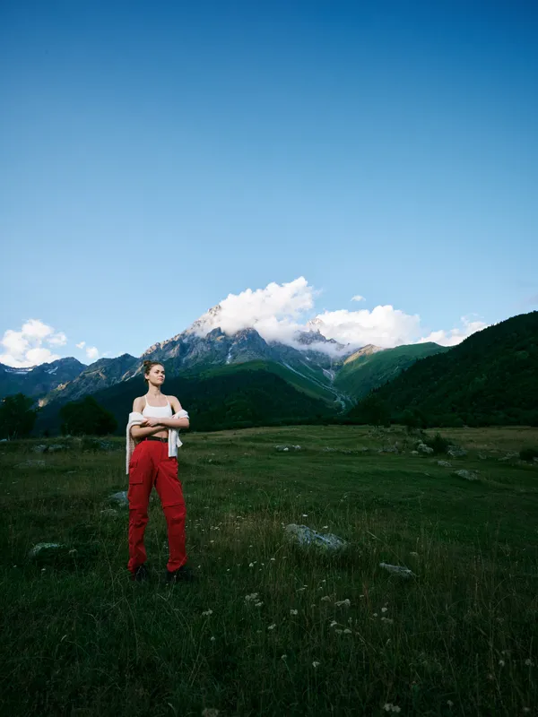Woman stands in a mountain meadow with bold redpants, under a clear blue sky, capturing outdoor adventure and serene nature amid alpine scenery